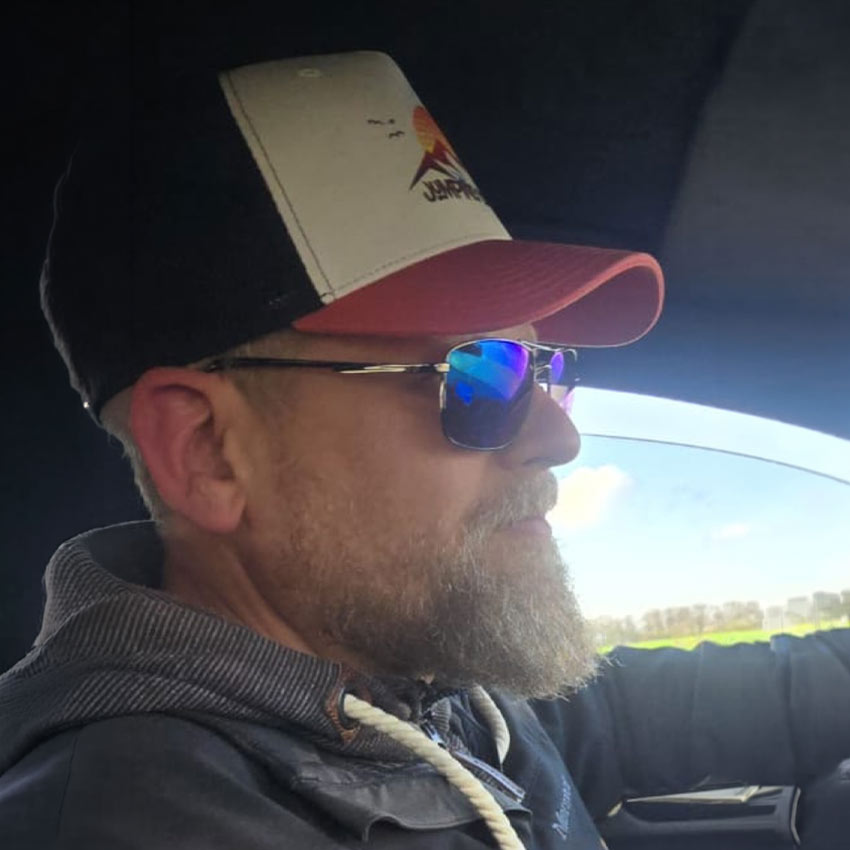 Ein Mann mit Bart und Brille fährt im Auto und trägt eine dunkle Jacke sowie eine Mütze