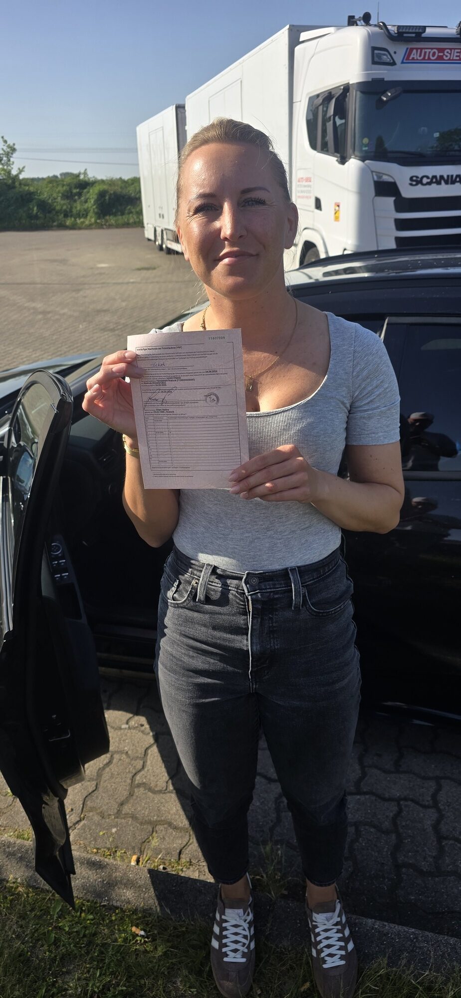 Frau mit leichtem Laecheln im grauen Shirt haelt Papier vor schwarzem Auto, weisser Lkw im Hintergrund