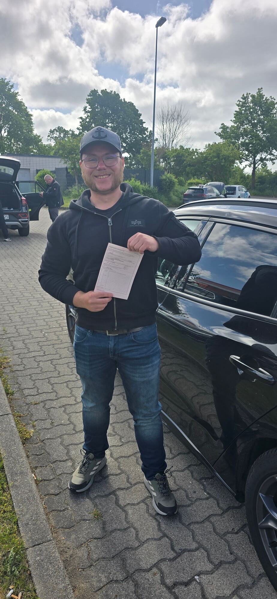 Laechelnder Mann mit Brille und schwarzer Kappe haelt Papier am Auto