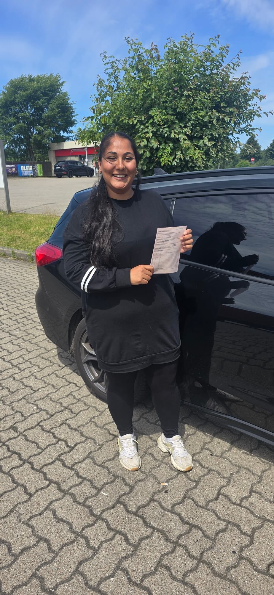 Laechelnde Frau in schwarzer Kleidung haelt Papier vor schwarzem Auto