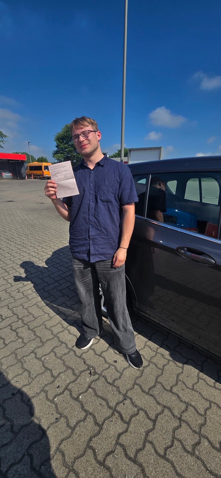 Lachender Mann mit Brille im blauen Hemd hält Papier neben schwarzem Auto