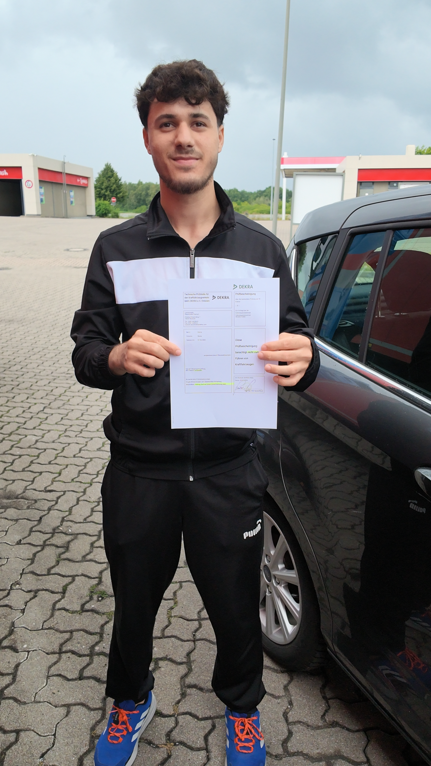 Laechelnder Mann im schwarzen Trainingsanzug haelt Dokument vor Auto