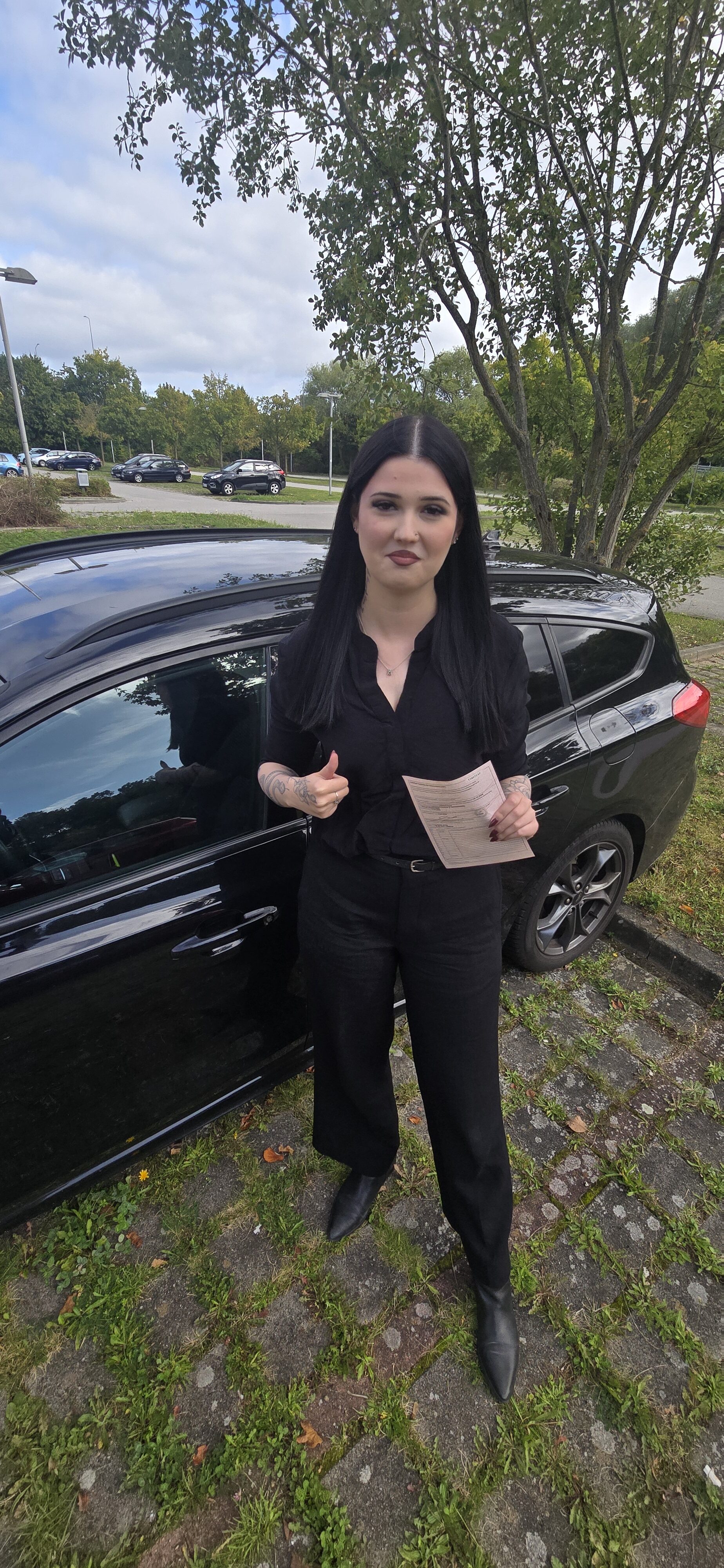 Junge Frau in Schwarz zeigt Daumen hoch mit Dokument vor schwarzem Auto