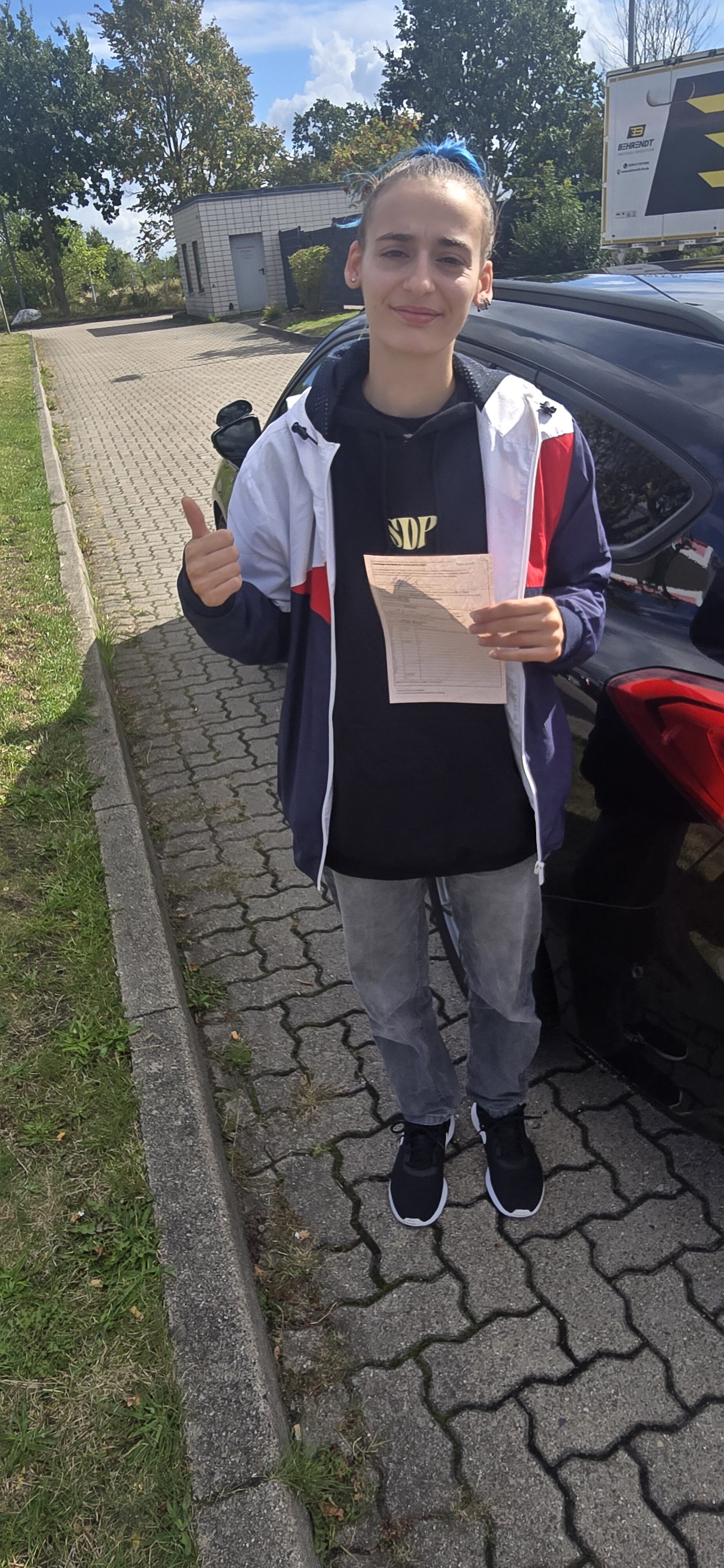 Junge Person mit blauen Haaren zeigt laechelnd Daumen hoch und haelt Dokument