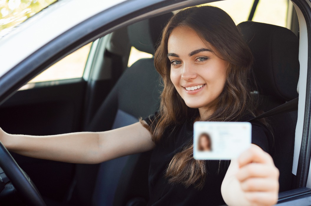 Ein glücklicher Fahrschüler sitzt in dem modernen silbernen Auto und zeigt der Kamera seinen Führerschein